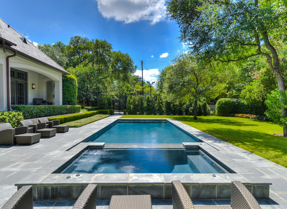 A modern outdoor space featuring a rectangular swimming pool bordered by gray stone tiles. Lounge chairs are positioned nearby on a patio, with a well-maintained lawn and trees in the background under a blue sky with fluffy clouds.