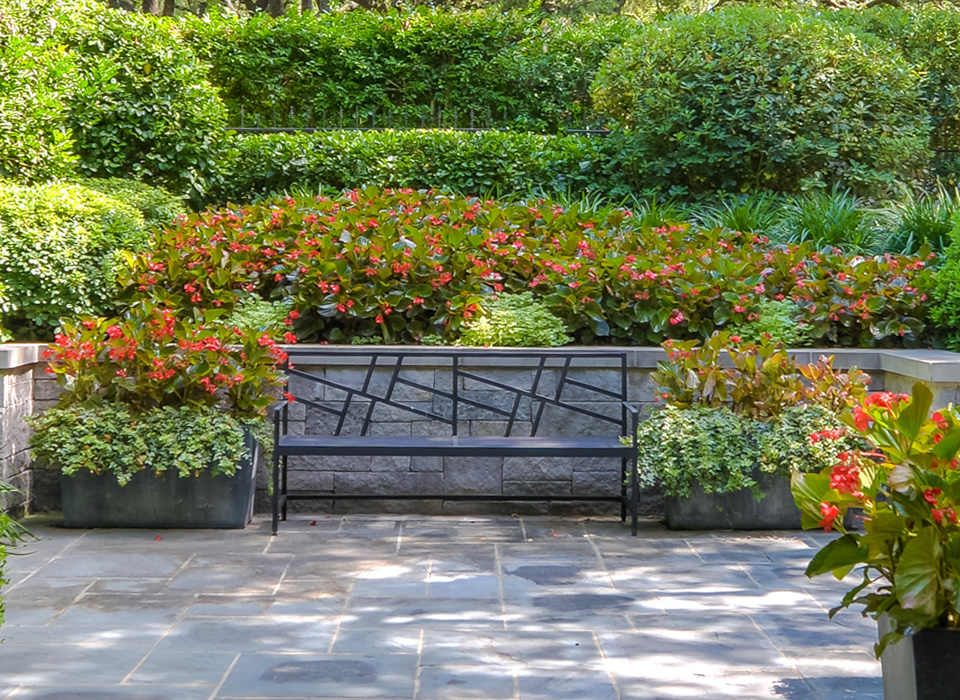 A metal bench in a landscaped garden surrounded by vibrant flower beds. The area features well-maintained greenery with varying heights of plants, creating a serene outdoor space.