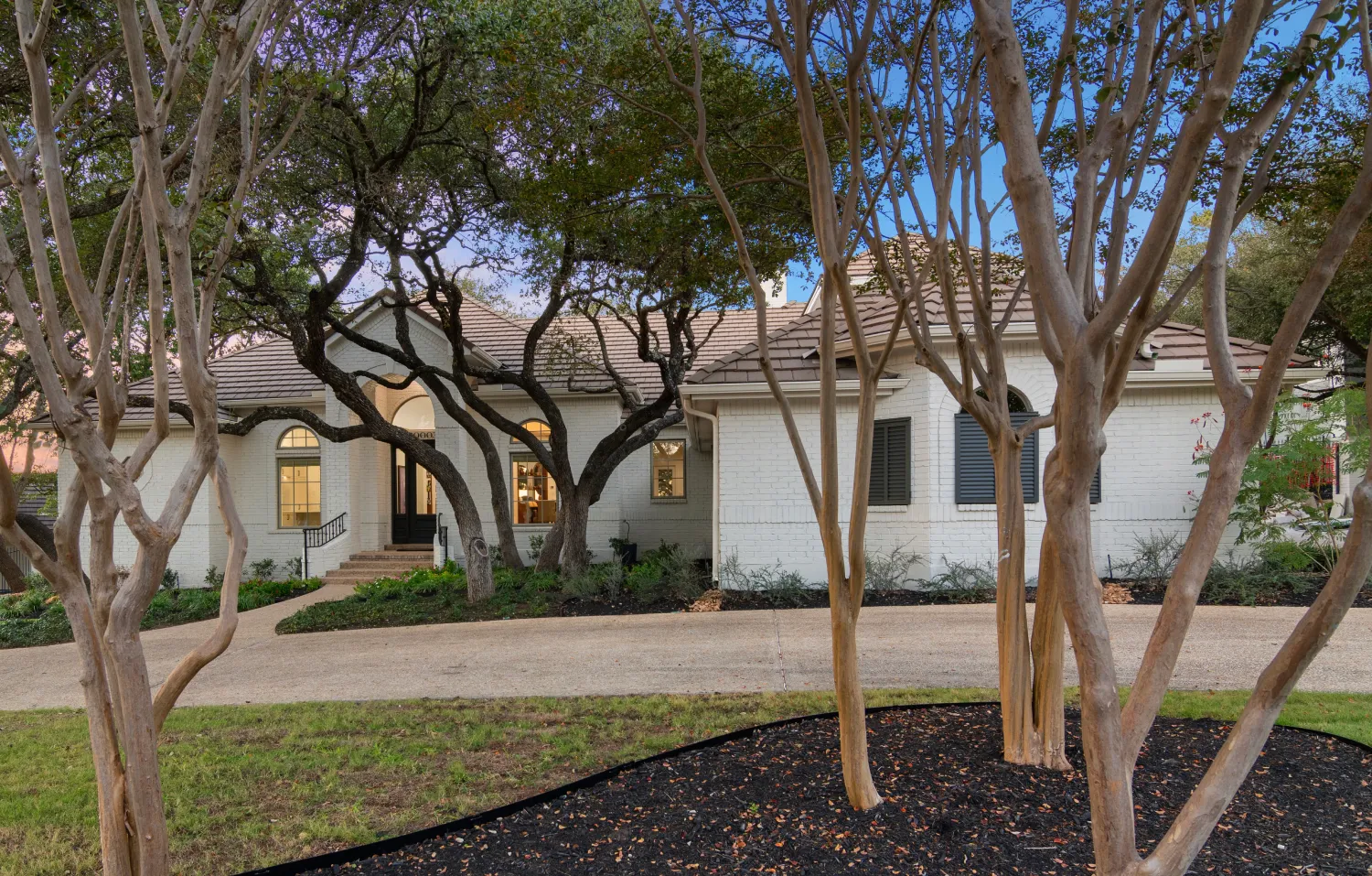 A single-story house with a light-colored facade, surrounded by trees. The entrance features a set of steps leading to a front door, while a curved driveway provides access to the home. The landscaping includes grass and flower beds.