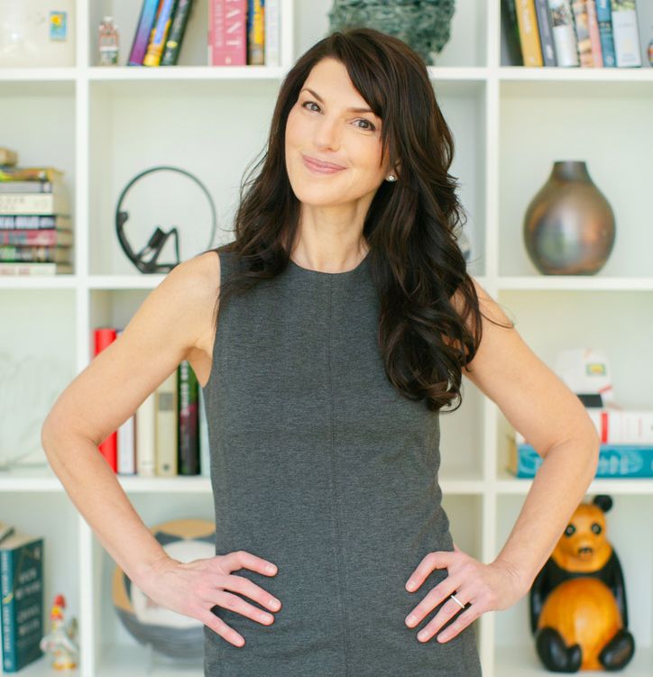 Deborah Miller from The Tesh Project:A woman with dark, wavy hair stands with her hands on her hips, wearing a gray sleeveless dress. Behind her is a white bookshelf filled with various books and decorative objects including a small ceramic bear.